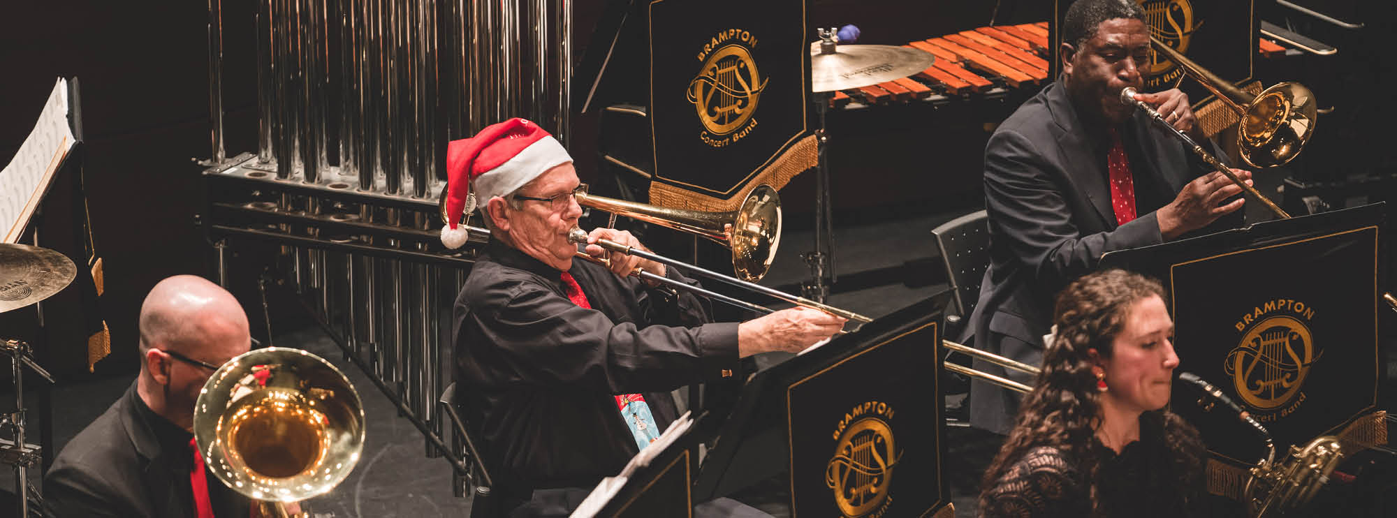 Image of 5 members of the Brampton Concert Band playing brass and woodwind instruments. They are all wearing red and black clothing, and two are wearing Santa hats. On each music stand hangs a banner bearing the name “Brampton Concert Band.” There are percussive instruments in the background. 