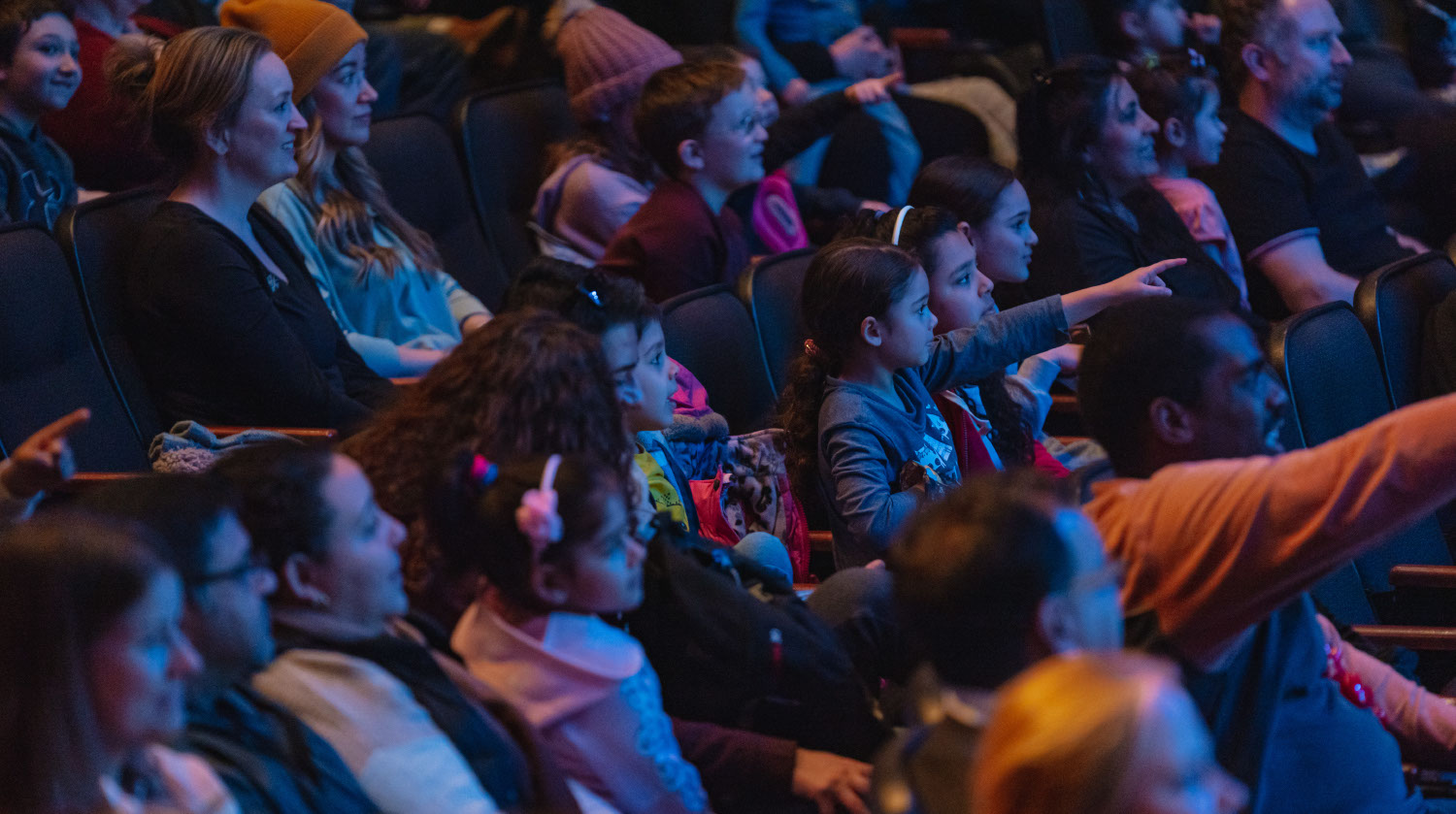 An image of a dimly lit theater with audiences of all ages seated, looking ahead and smiling. One child points forward.