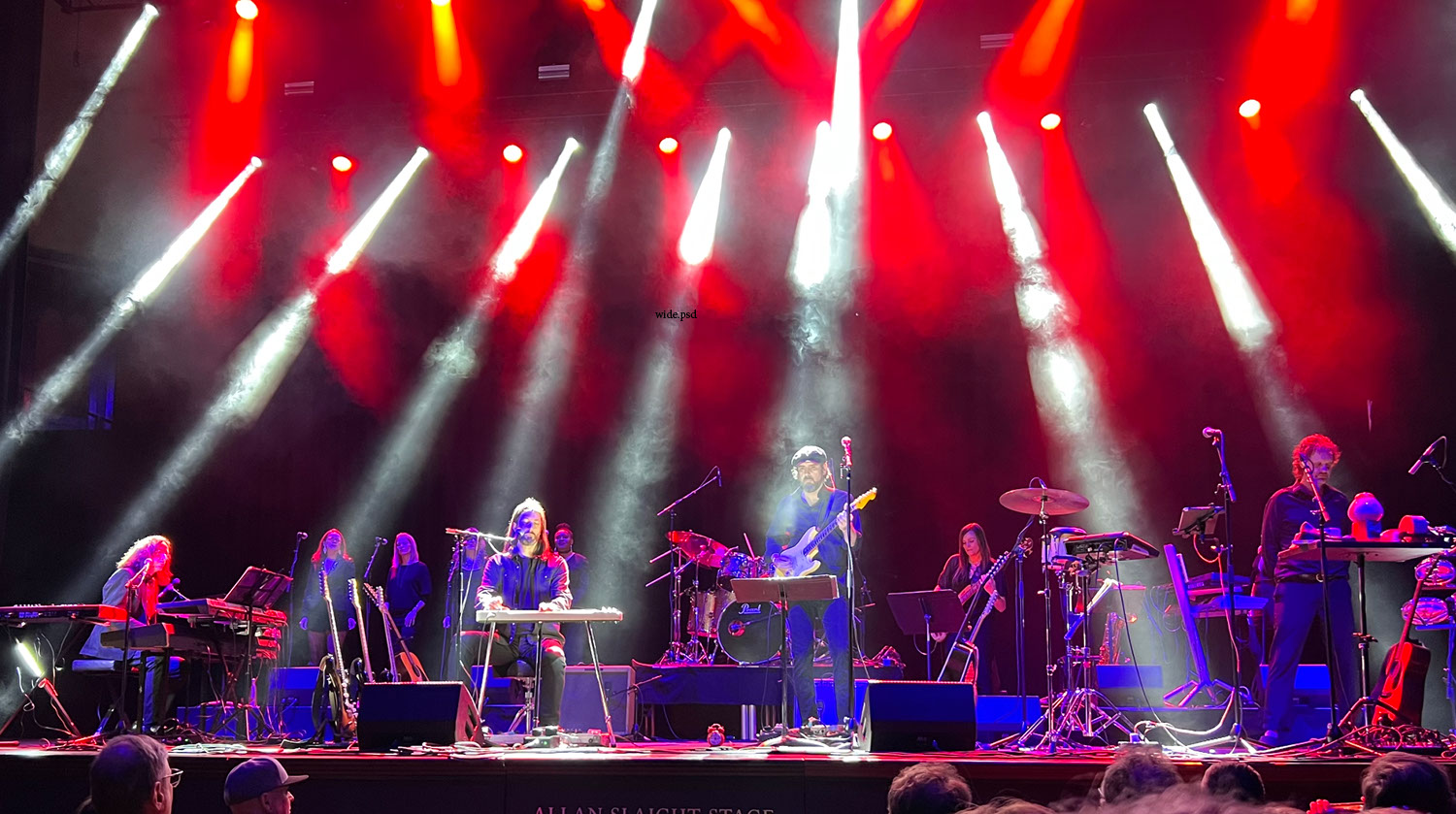 Image of several musicians performing on stage. White rays of light cut through smoke, which is backlit in red. The artists on stage are highlighted in shades of deep blue and red. They play an array of instruments including guitars, the piano, drums and a keyboard. Several music stands, microphones and cables are splayed across the stage. In front of the stage, the backs of people watching the performance can be seen. 