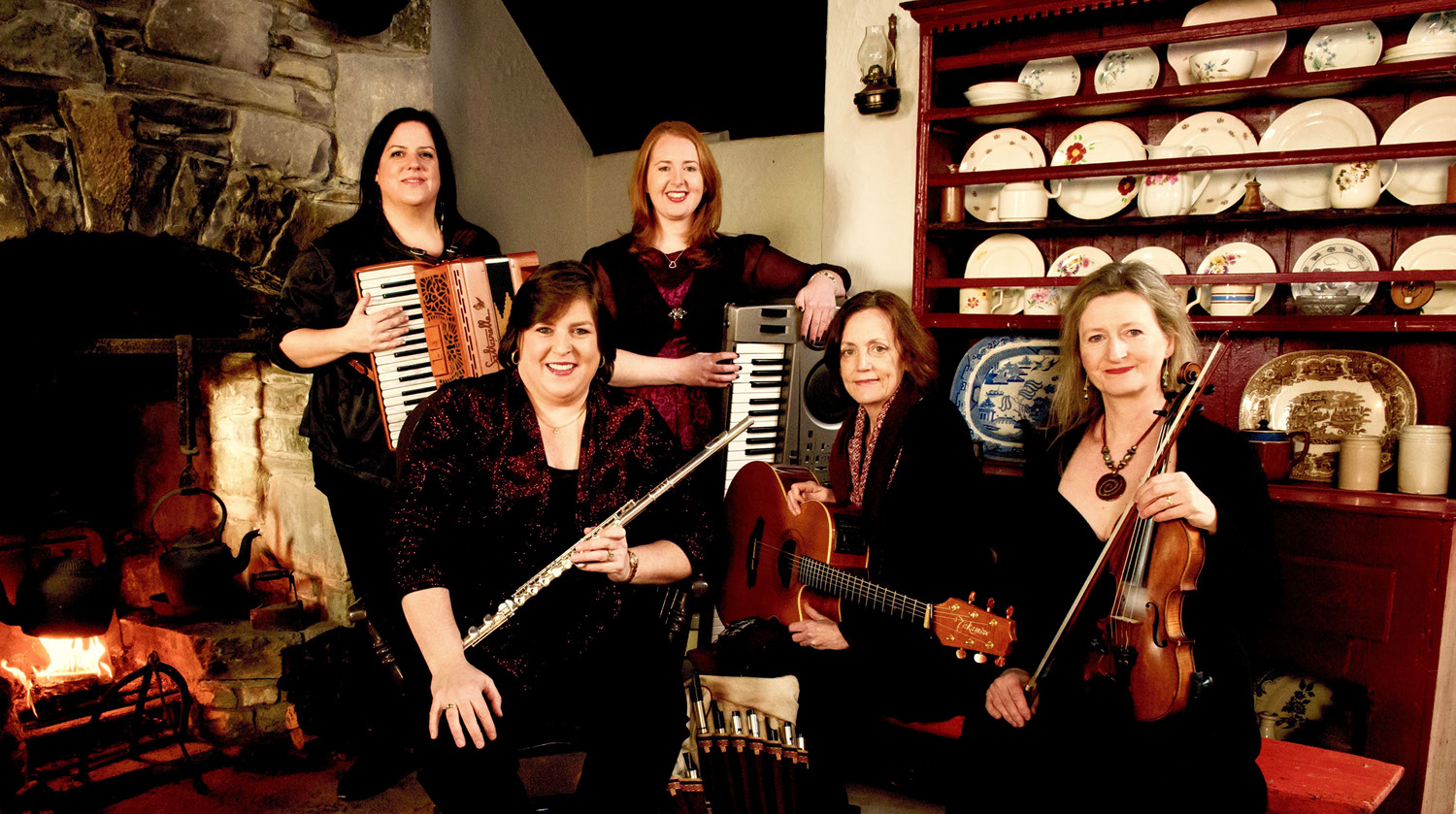 A posed image of the five women that comprise Cherish The Ladies the band. The woman hold an array of instruments, including an accordion, flute, acoustic guitar, violin and keyboard. They are smiling and wearing dark, muted colours. Two of the women stand, and the other three sit in front of them in an old home. To the left, a wood fire is burning within a stone mantel. On the other side of the room, white china is displayed on a tall wooden credenza with several shelves.