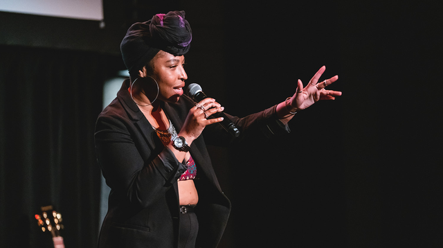 Image of curator The Wild Woman holding a microphone up to her mouth with her other arm outstretched as she performs spoken word poetry. She has deep skin and her hair is up in a black head wrap knotted at the front. She is wearing a bralette top under a black suit with silver jewellery. In the background there are black curtains, the bottom corner of a projector screen, and a guitar propped up. 