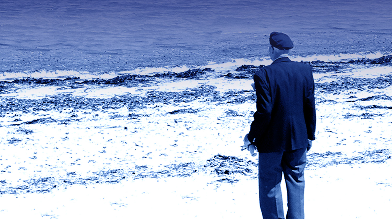 Image of an older man with white hair, likely a veteran, facing away toward a shoreline. He is wearing a dark suit with an old-fashioned cap. He’s also holding something in his left hand. The sandy beach meeting the water is in the background. 