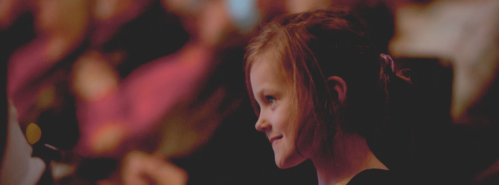 Little girl in an audience, she is wearing a black dress, has blonde hair and similing. This image is for Giving Tuesday