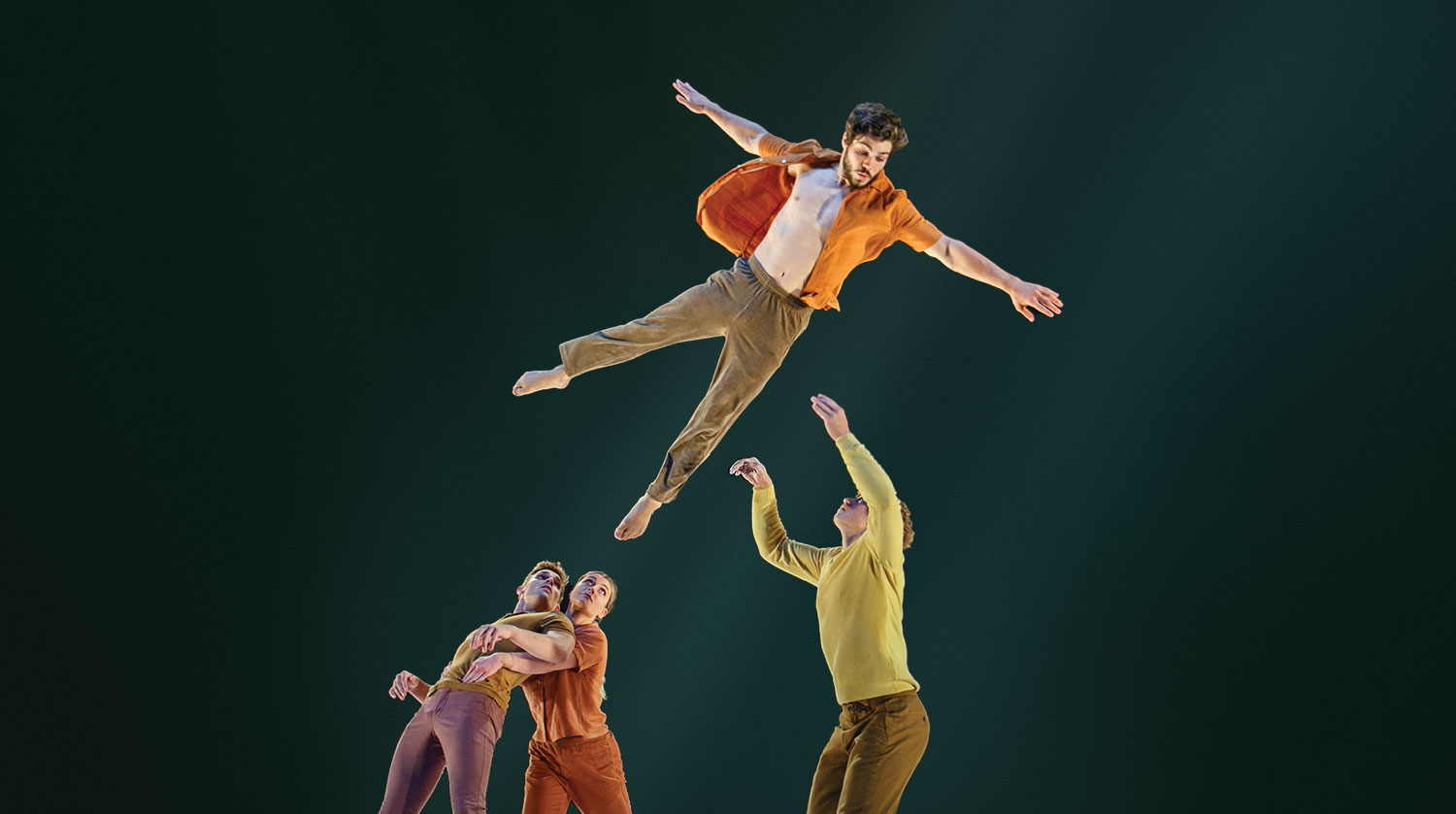 Image of three men and one woman wearing neutral clothing amidst a black backdrop while performing a circus stunt. The man standing to the right wears a yellow shirt and brown pants, and has his arms splayed upwards into the air. Another man is propelled in the air above him, wearing an open orange shirt, with his arms and legs spread wide. Beneath him on the floor, a man and woman embrace one another. He leans his weight into the arms of the woman, who holds him and looks up above her. 