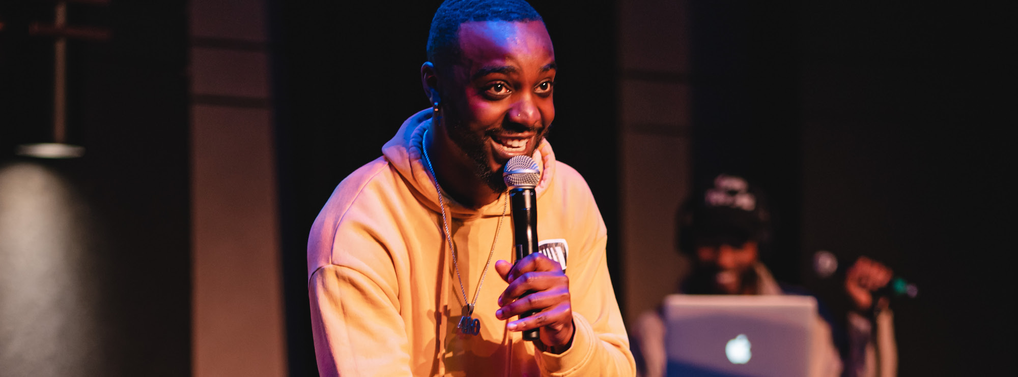 Image of curator Jemane Kent leaning on his knee while holding a microphone up to his mouth and smiling. He has dark skin, and dark hair and facial hair. He’s wearing a yellow hoodie and a long silver necklace. Behind him, there is a man using audio equipment and a silver laptop. There is a grey and black wall with a lit wall sconce in the background. 