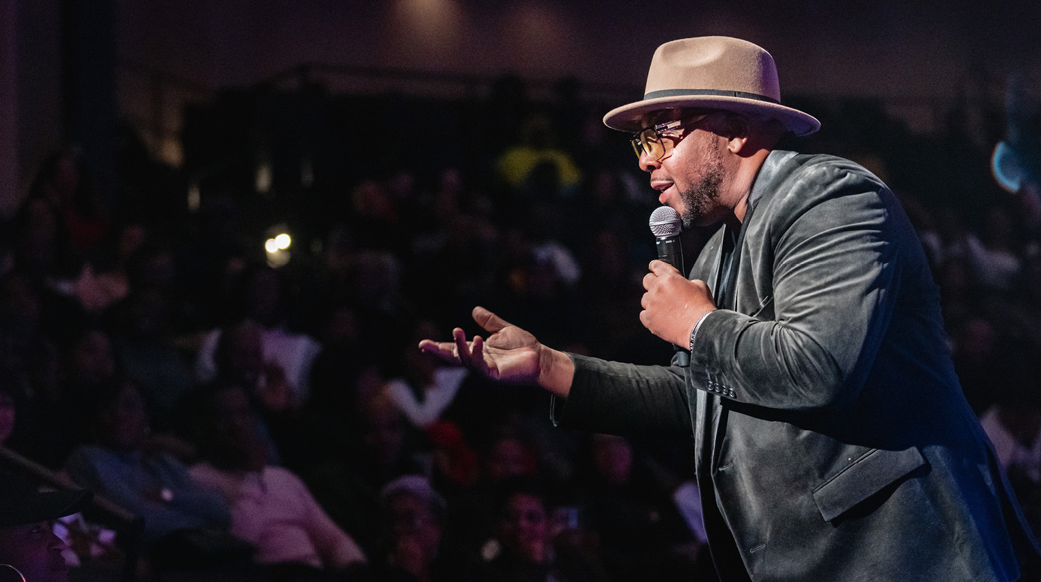 Image of comedian and curator Jay Martin leans toward the audience with a hand outstretched, while holding a microphone up to his mouth as he says something to the crowd. He has deep skin and dark hair and some facial hair. He’s wearing a black suit and brown brimmed hat with glasses. The large audience is blurry in the background. 