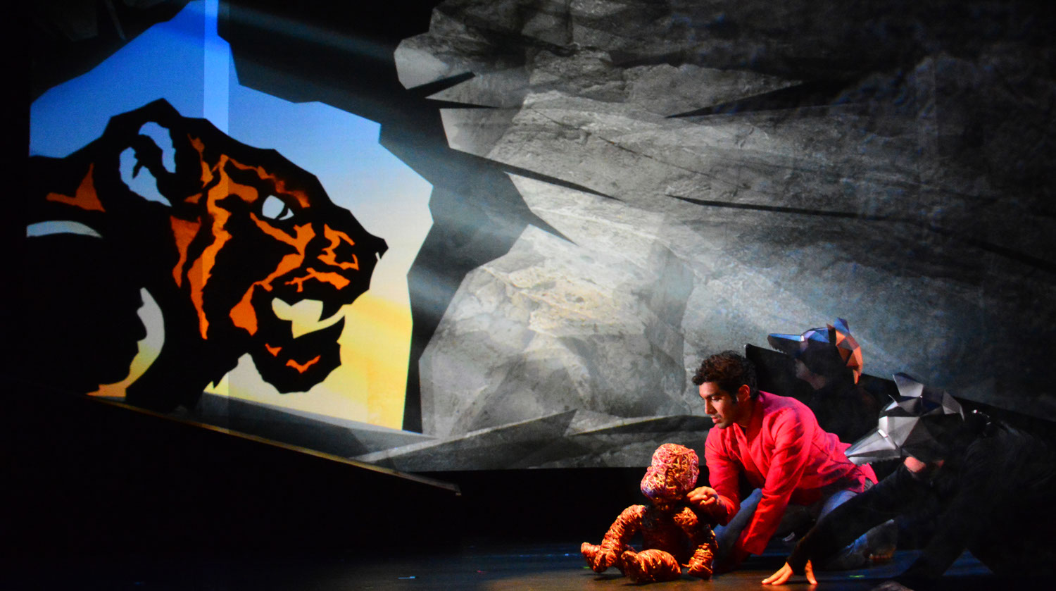 Image of stage which features a colourful, illuminated graphic of a tiger shining brightly over the backdrop of a stone wall. In the right corner of the stage, a man wearing a red shirt kneels on the floor beside other actors who are dressed in geometric wolf heads and black bodysuits. They all hover together, facing the direction of the tiger before them. 