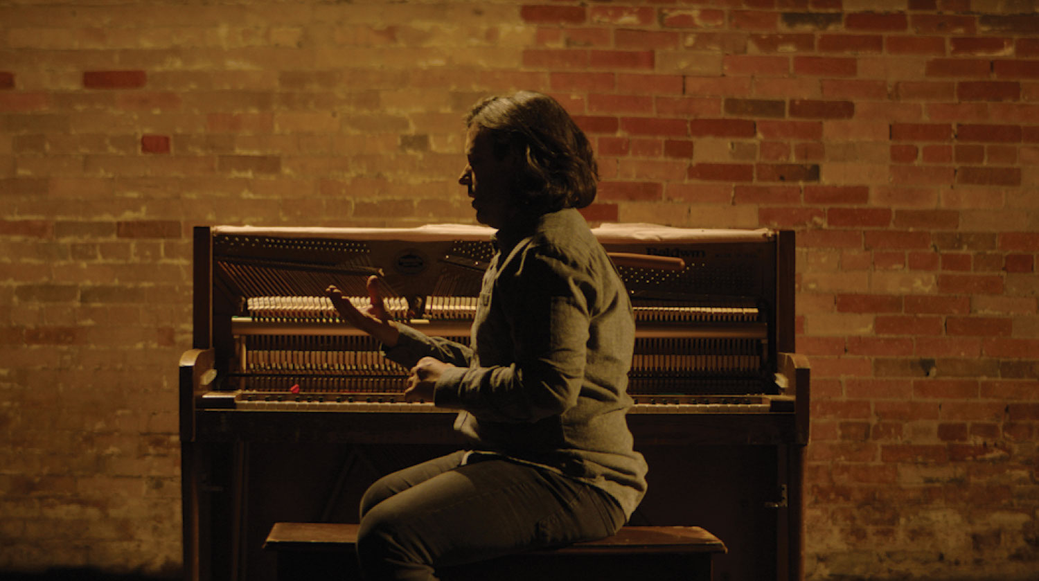 Actor James Smith sits on a bench in neutral-coloured clothing before an antique piano. The wall behind the piano features exposed red bricks. He puts his hands out in front of him to look at them. The room he is in is dimly lit and casts a shadow over his body. 