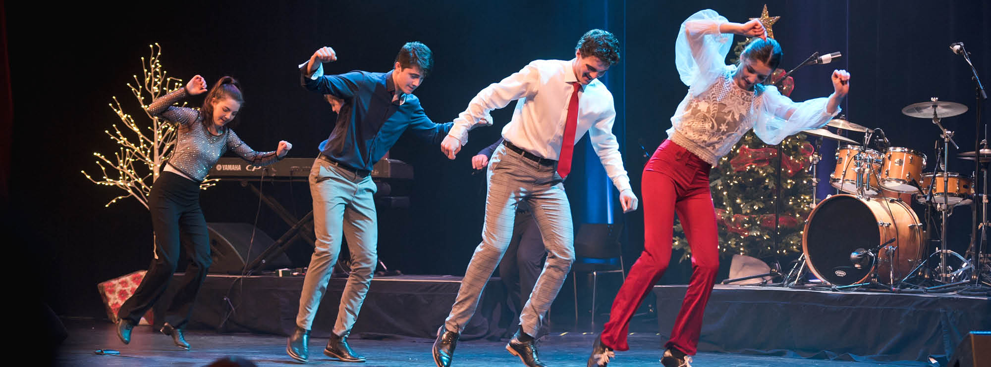 Image of four members of The Next Generation Leahy step-dancing on stage. Two are girls and two are boys, and they all have light skin and dark hair. There are instruments and Christmas decorations in the background. 