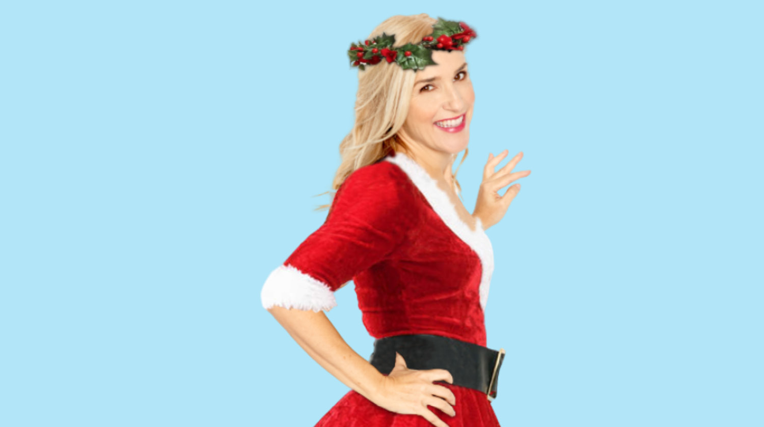 Image of actress and comedian Anne Marie Scheffler smiling and wearing a red velvet Santa-style dress with white fluffy trim, along with a holly and ivy crown. She has light skin and blonde hair. The background is light blue.    