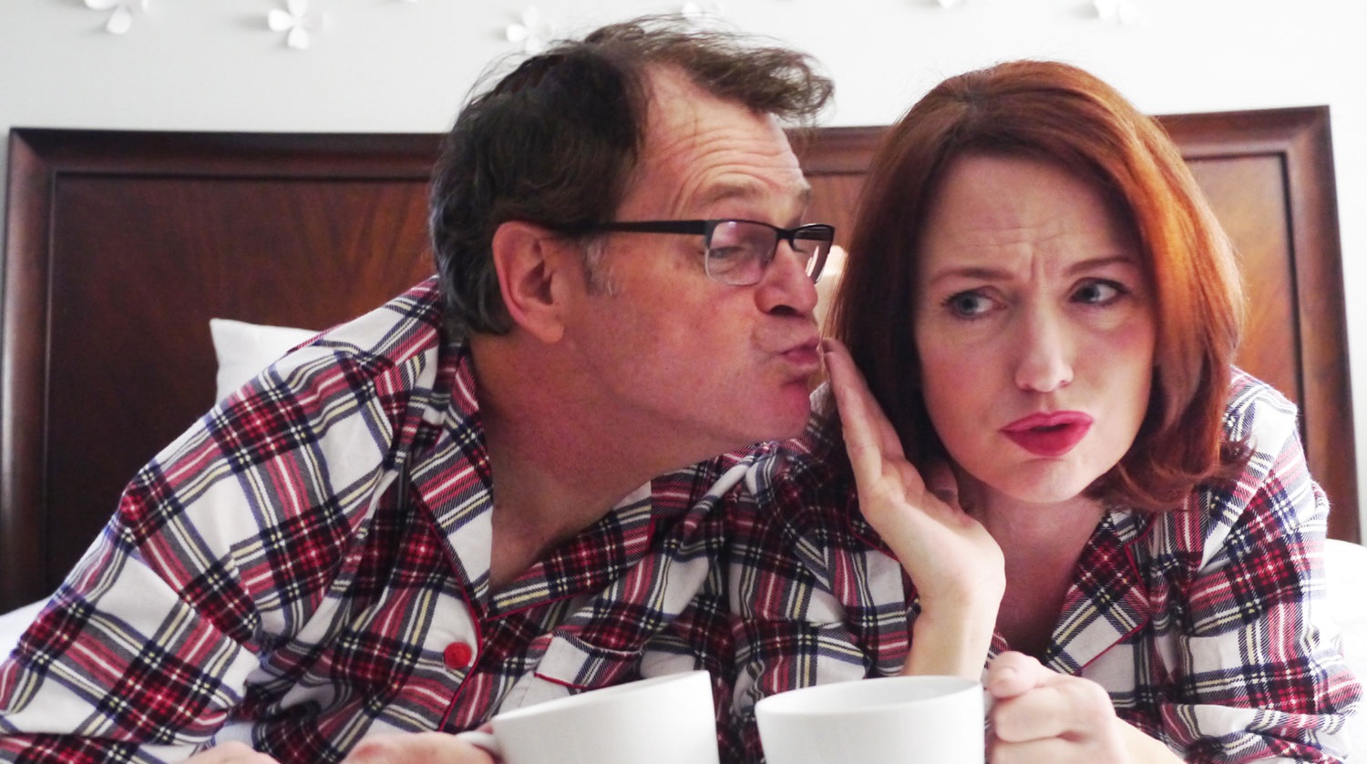 Image of a man and woman dressed in matching red and white flannel pajamas. They both lay side by side on their stomachs atop a bed, which has a wood headboard. They hold white mugs in their hands. The man wears glasses. He is trying to kiss the woman beside him, but she puts her hand up between his lips and her cheek. 