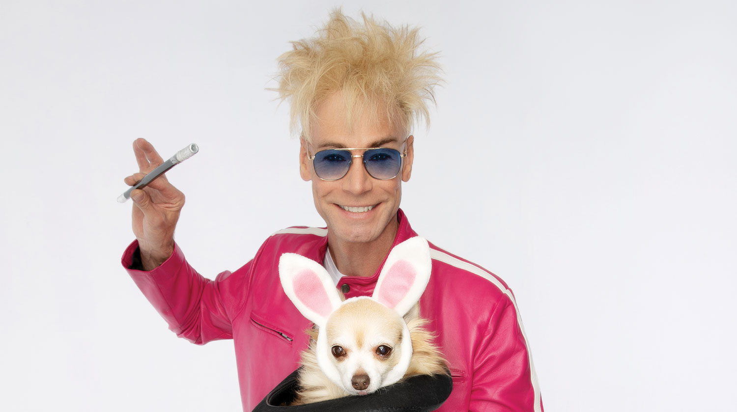 Image of Murray The Magician smiling before a white backdrop. He has light-toned skin and blonde teased hair. Murray has a wand raised in his right hand. He wears aviator sunglasses with blue lenses, and a pink jacket with a white stripe across the shoulders. In his left hand, he holds a black hat. Inside of the black hat, there is a white fluffy dog wearing bunny ears.