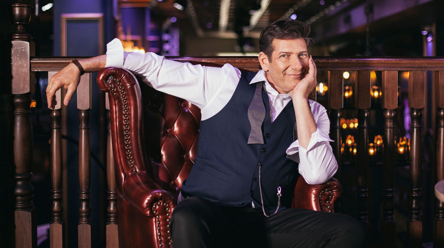 Image of singer-songwriter Micah Barnes sitting on a brown leather chair before a wood banister in a dimly-lit room. He has pale-toned skin, blue eyes, and brown hair. He wears a white dress shirt and a navy blue vest atop it, with an undone grey bowtie draped around his collar. He leans his right arm over the back of the chair and his left arm on the armrest as he props up his head in his hand.
