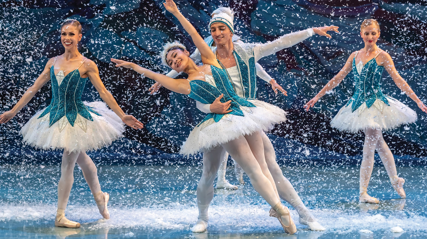 Image of dancers from Canada’s Jörgen Ballet performing in The Nutcracker. There are three ballerinas and two male ballet dancers dressed in blue, silver and white costumes dancing while faux snow falls around them. 