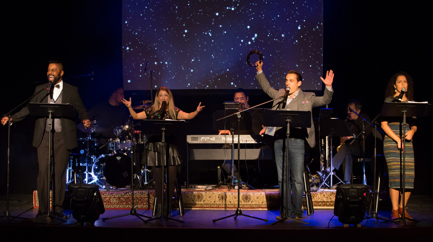 Image of four singers and three musicians performing on stage in front of a blue night sky projection. The singers, two male and two female, stand downstage with music stands and microphones in front of them. Behind them, the male musicians play drums, keyboard, and saxophone. 