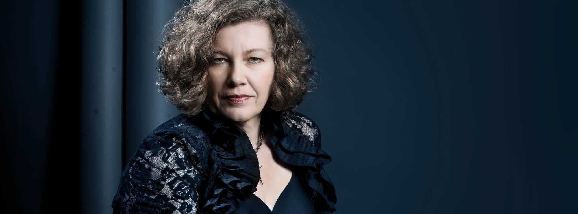 Image of a Canadian vocalist Adi Braun dressed in all-black against a dark background. She has light skin and greying curly shoulder-length hair. 