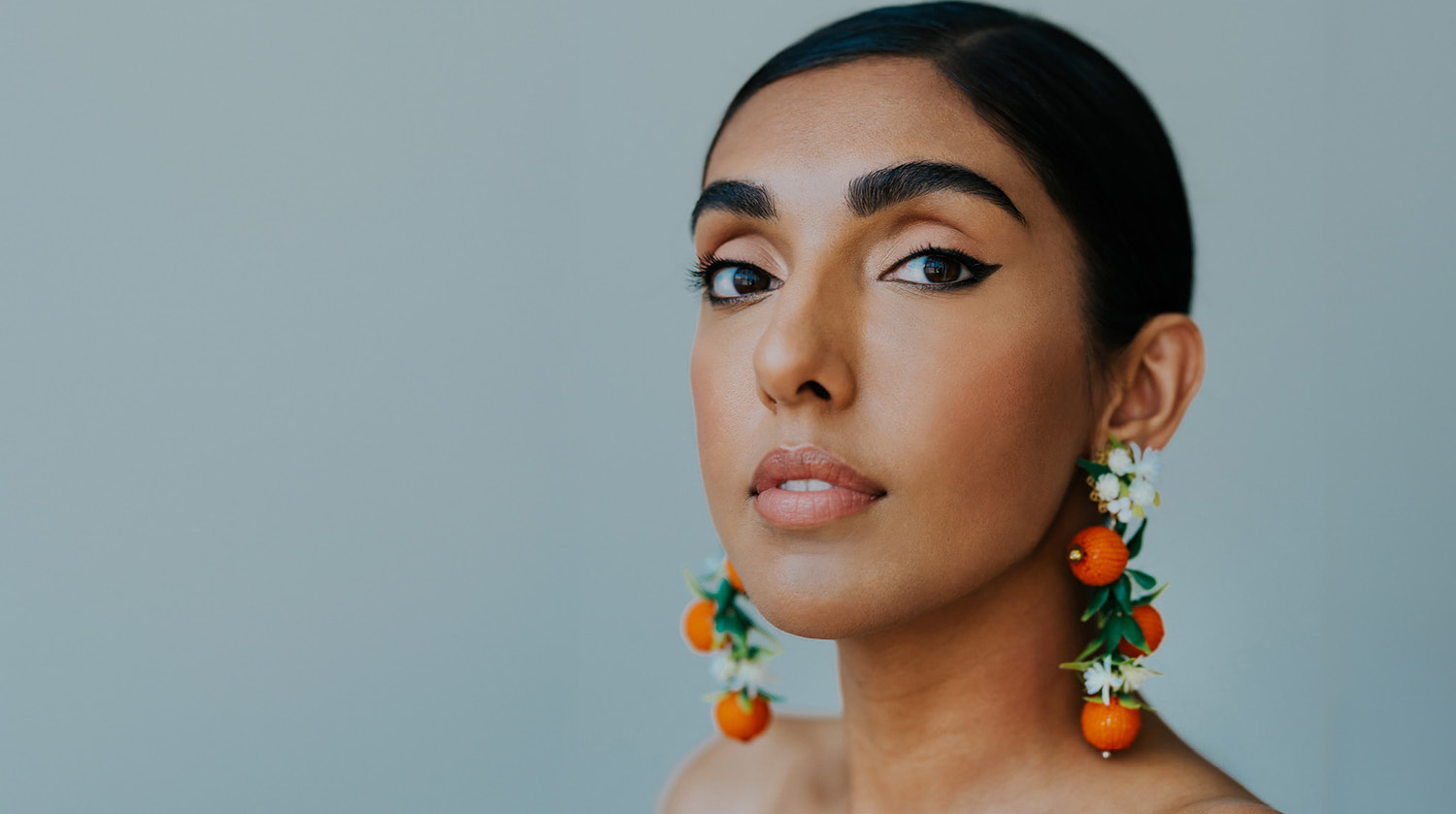 Image of Rupi Kaur before a pale blue-grey backdrop. She has warm-toned skin, deep brown eyes, and slicked-back black hair. She wears long beaded earrings, adorned with oranges and white flowers. 
