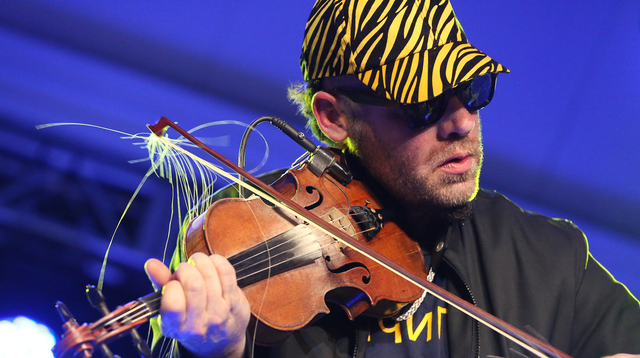 Image of Canadian fiddler Ashley MacIsaac playing the fiddle with a frayed bow. He has light skin and brown hair with light facial hair. He’s wearing a black and yellow striped baseball cap, with black sunglasses, and a black and yellow shirt and jacket. The background is blue. 