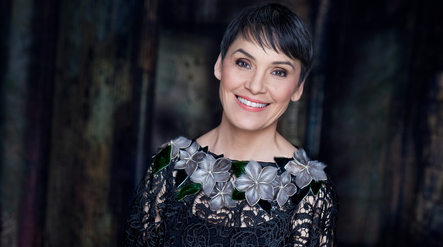 Image of Inuk singer Susan Aglukark smiling, wearing a black lace dress with grey flowers around the neckline. She has light skin, brown eyes, and short pixie cut black hair. 