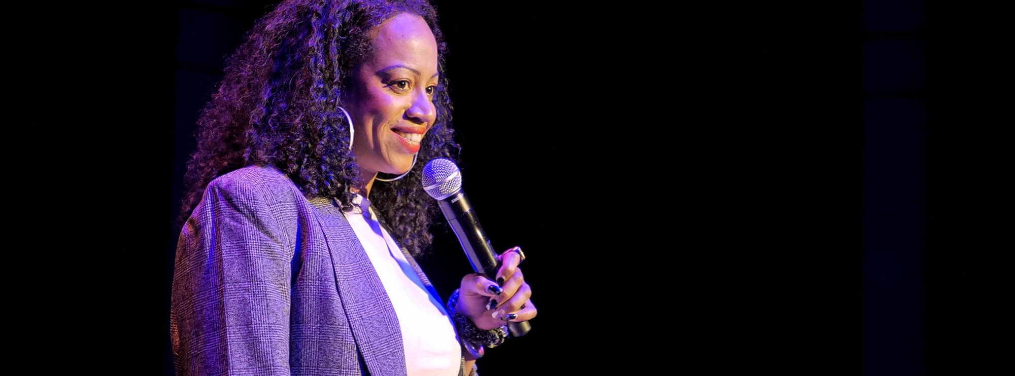 Image of curator Keesha Brownie with deep skin and dark wavy hair holding a microphone up to her mouth and smiling. She is wearing a white t-shirt with a grey plaid blazer and some jewellery. 