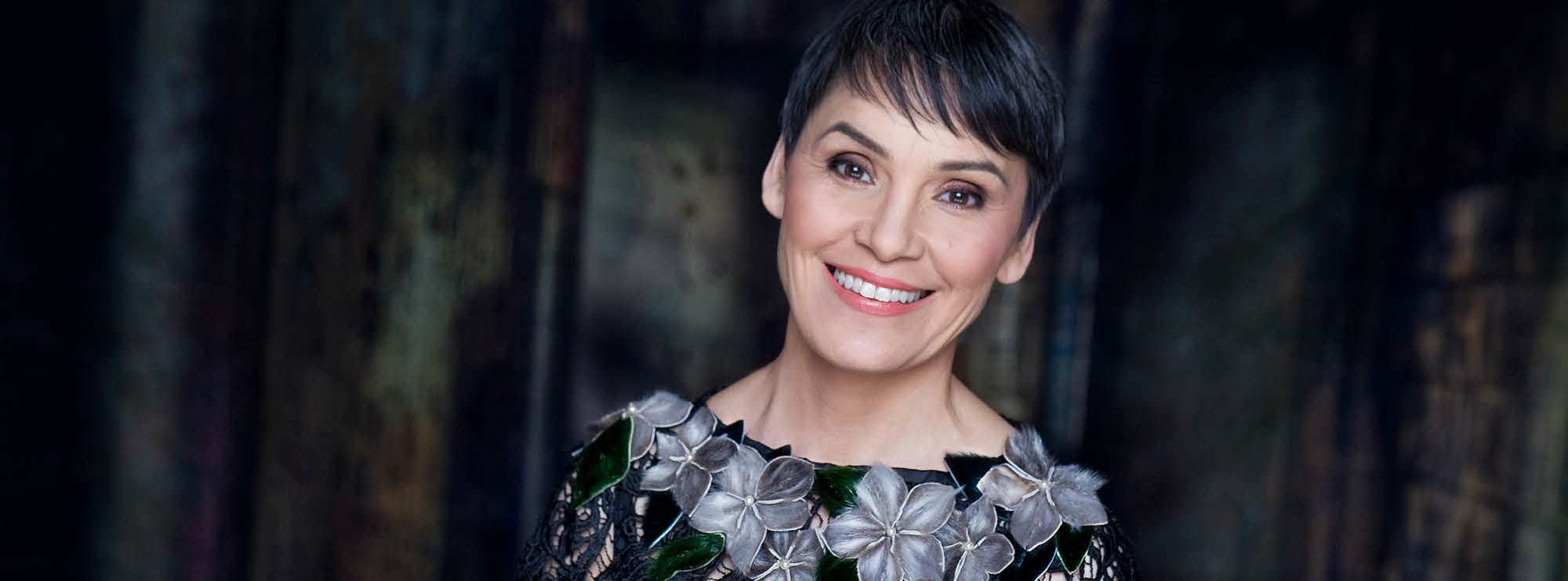Image of Inuk singer Susan Aglukark smiling, wearing a black lace dress with grey flowers around the neckline. She has light skin, brown eyes, and short pixie cut black hair. 