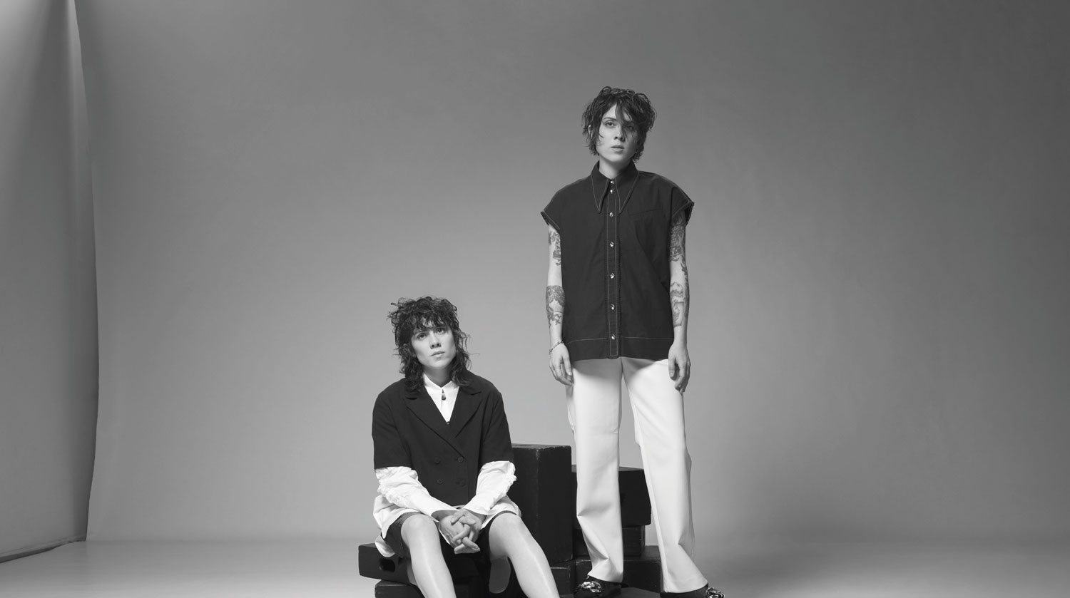 A black and white image of Tegan and Sara before a plain backdrop. The woman to the left is seated atop two black boxes. She has light-toned skin and shoulder-length curly hair with bangs. She wears a black jacket with buttons, on top of a white long-sleeve shirt. The other woman stands to the right of her. She has pale-toned skin and short hair. She wears a black vest, white pants and has several tattoos on both of her arms.