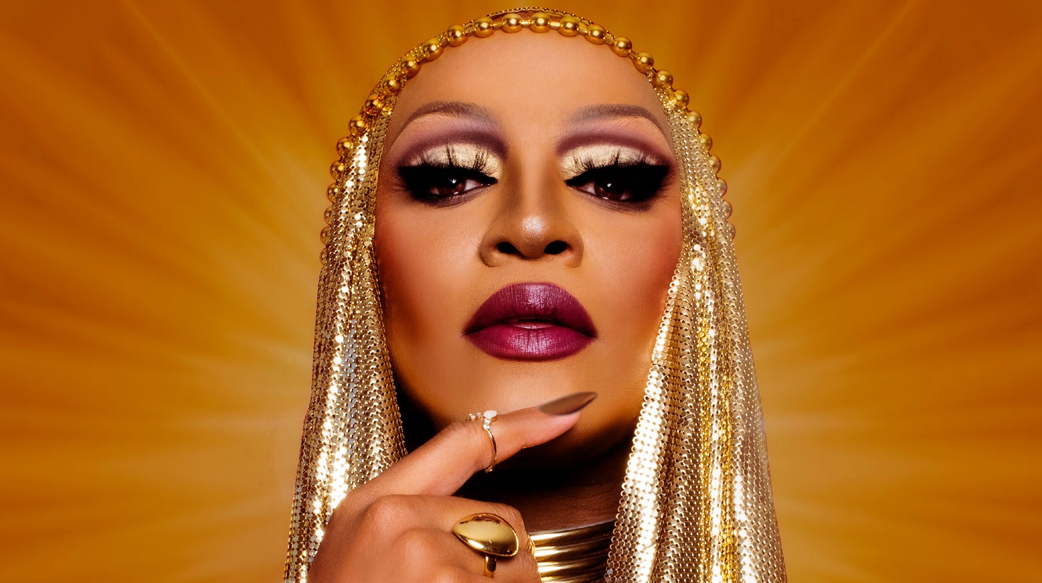 Image of Tynomi Banks. Her face is tilted upwards and her right hand is placed against her chin. She has deep-toned skin. Her nails are painted brown and she wears two gold rings. She wears an elaborate, gold beaded headpiece and thick necklace. Her eyes are outlined with gold eyeshadow and black liner. She wears dark magenta lipstick and face makeup. Rays of gold and brown light shine from behind her head.