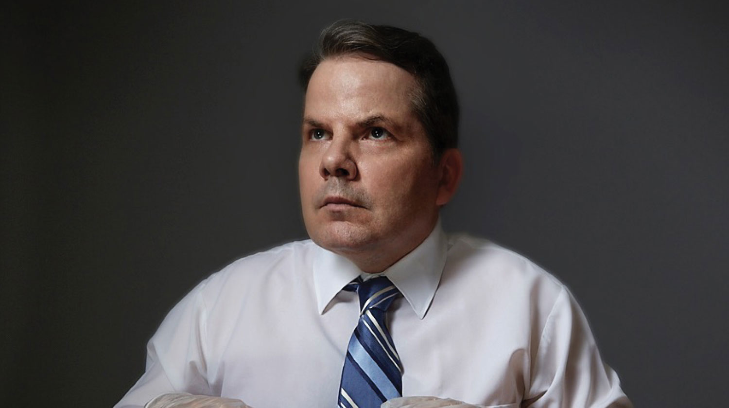Image of Bruce McCulloch before a dark grey background. He has light-toned skin, brown eyes and brown hair. He looks upward with a serious expression on his face. He wears a white dress shirt with a blue stripped tie. 