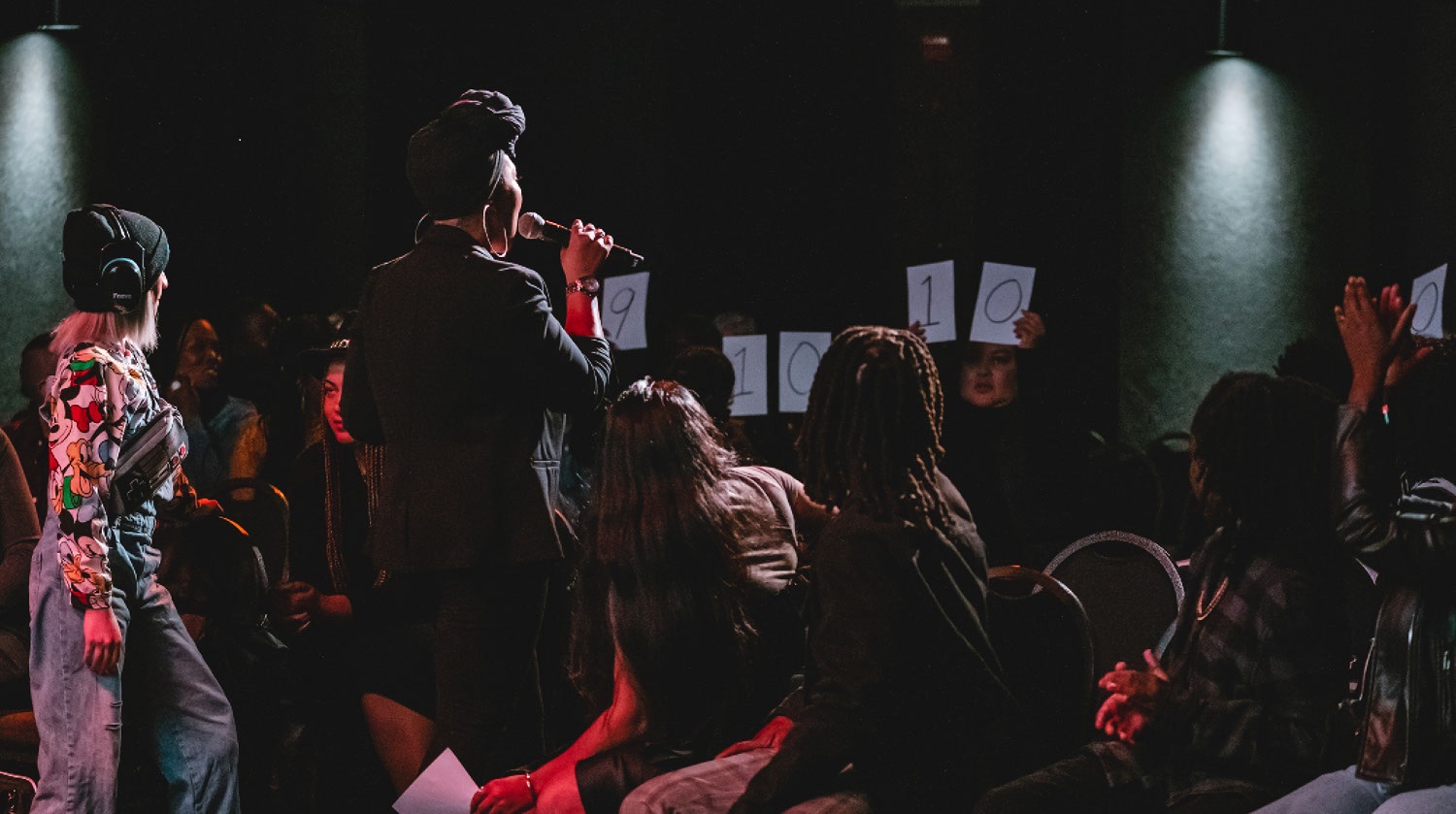 Image of a dimly lit room full of people. Two people can be seen from behind standing. One of the women that is standing speaks into a microphone. She is dressed in all black clothing with a black headwrap and large hoop earrings. The people seated around her turn to face the back of the room. The seated individuals at the back of the room hold white pieces of paper above them. The papers display an array of black numbers written on them. 