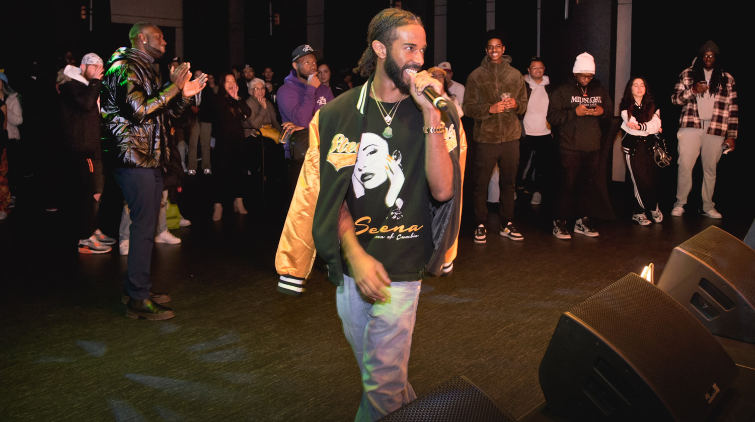 Image of curator and rapper TCspades holding a microphone up to his mouth and smiling while looking toward the stage. He has deep skin, dark braids and facial hair. He’s wearing a black and gold letterman-style jacket and a t-shirt with the singer Selena on it, along with two gold necklaces and a watch. Behind him the crowd watches the stage with him. 