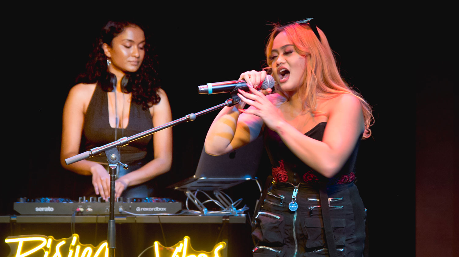 Image of two young women performing. The woman to the left has medium-toned skin and wears a black tank top. Around her neck is a set of headphones. She manipulates the turntable below her with her hands. The woman to the right has light-toned skin and orange hair. She wears a black tank top with red embroidery. In front of her is a microphone on a stand. She wraps her hands around the microphone and sings into it. 