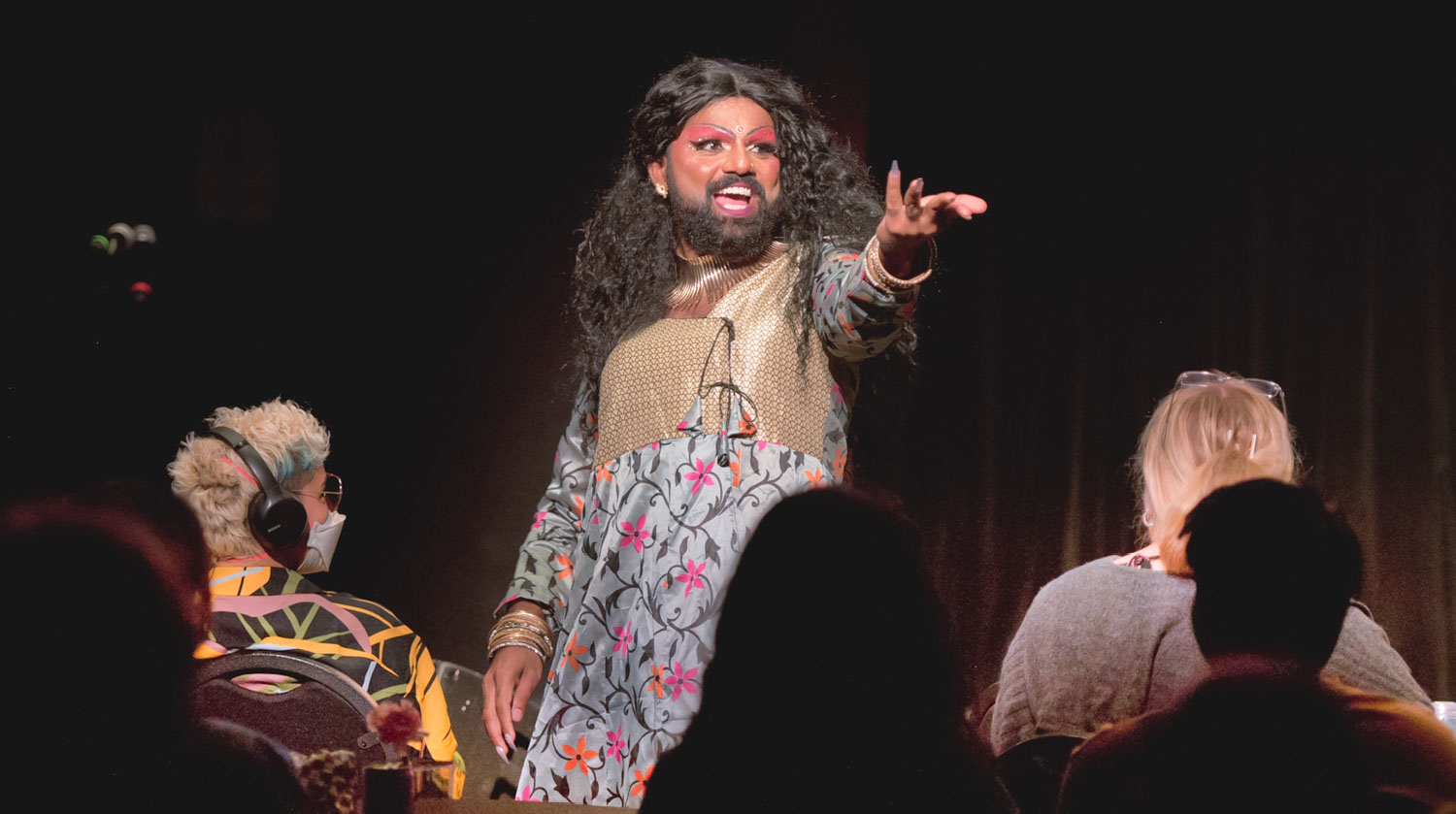 Image of deep-toned drag artist performing before an audience. The artist has long black hair and a beard. They are wearing bright pink eye shadow and decorative jewels around their face. The artist is outfitted in a brightly coloured dress with flowers embroidered on it. The top of the dress features ornate gold detailing. The artist smiles and reaches their hand out to the audience. Audience members can be seen sitting on chairs from behind, experiencing the performance. An audience member to the left has short hair with coloured streaks in it, and wears a vibrant patterned t-shirt with headphones atop their head.