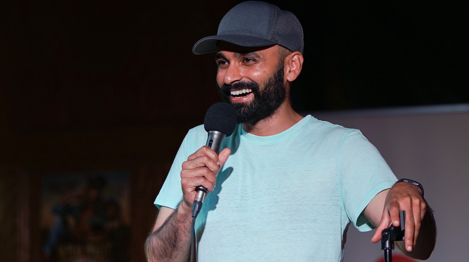 Image of curator Sunny Chahal holding a microphone up to his mouth and smiling. He has medium skin, dark hair and facial hair. He’s wearing a black baseball cap, a light blue t-shirt. He’s also wearing a watch on his left wrist and holding onto a microphone stand. The background is gray and black. 
