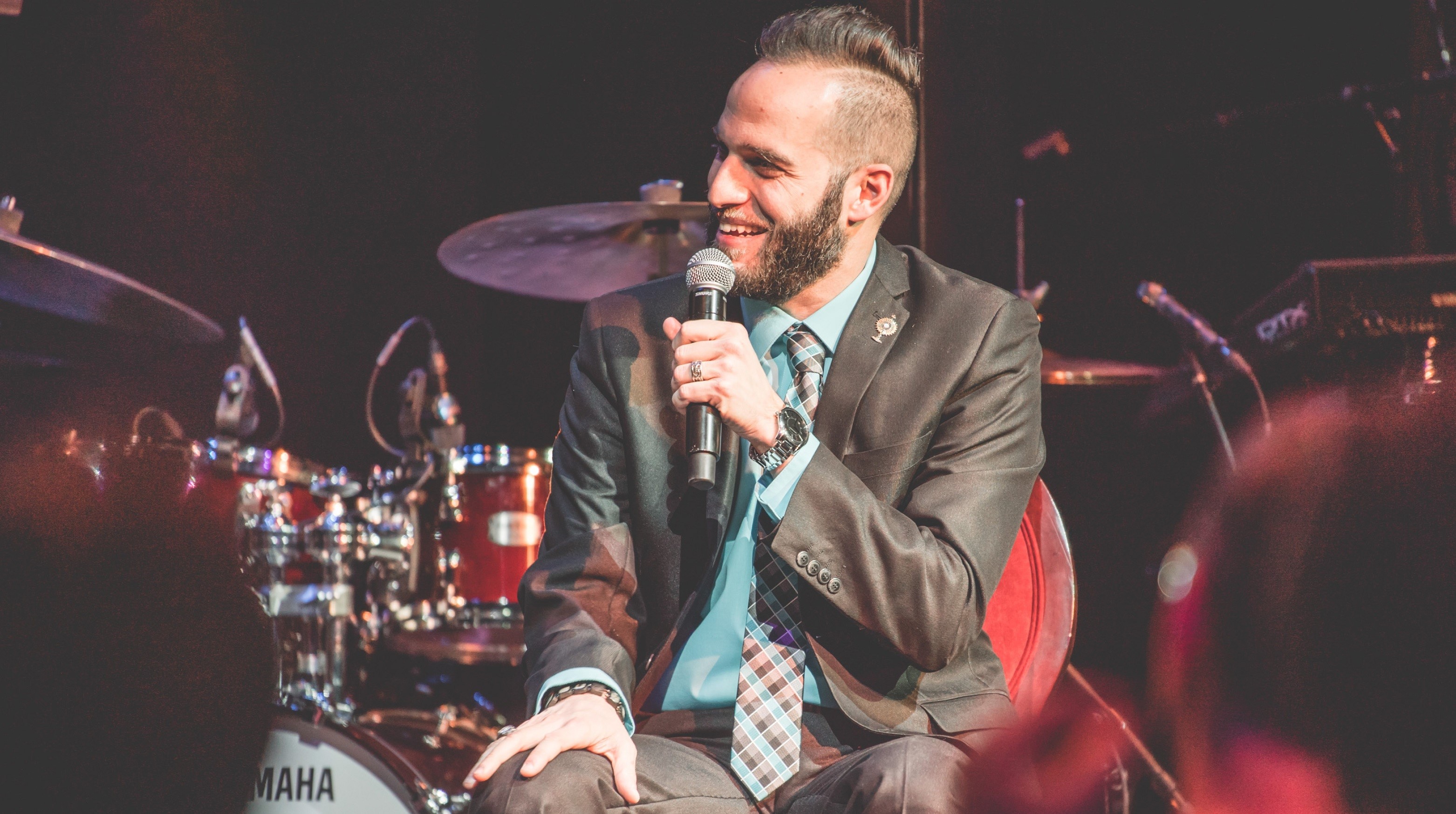 Image of curator Carmen Spada, with light skin and dark hair, holding a microphone up to his mouth and smiling. He is seated in front of a red drum set and other music equipment, and wearing a grey suit with a teal shirt. 