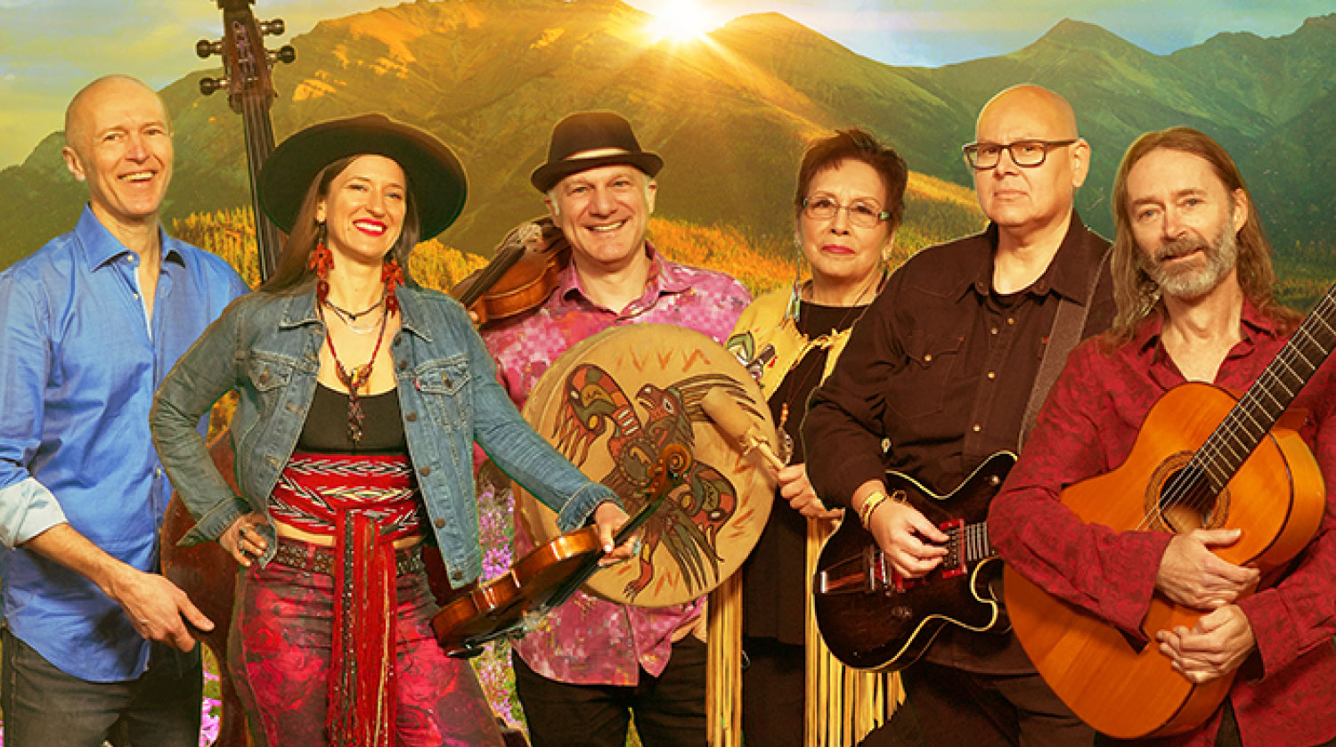 Image of the six members that comprise of Walking Through Fire the band. The musicians stand side by side smiling, wearing bright coloured clothing, and holding an array of instruments including an acoustic guitar, electric guitar, violin and hand-held drum. The drum displays an illustration of an eagle. Behind them is an image of a mountain with the sun setting beyond it.