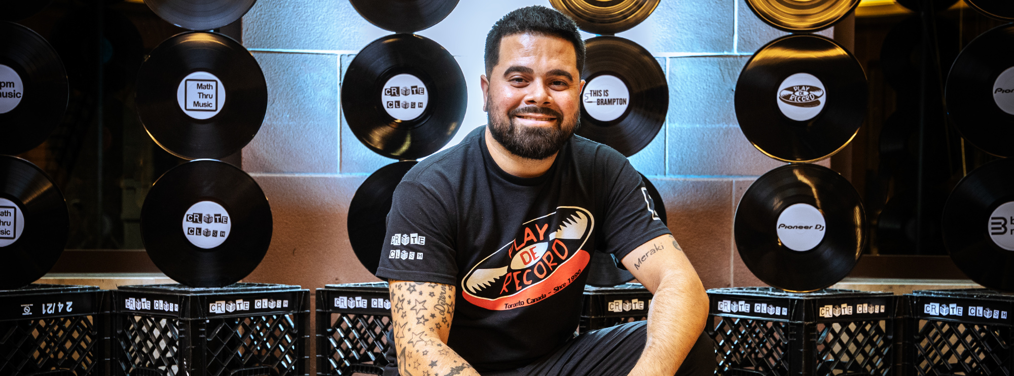 Man sitting and smiling in front of a wall decorated with vinyl records, wearing a "Play De Record" shirt.