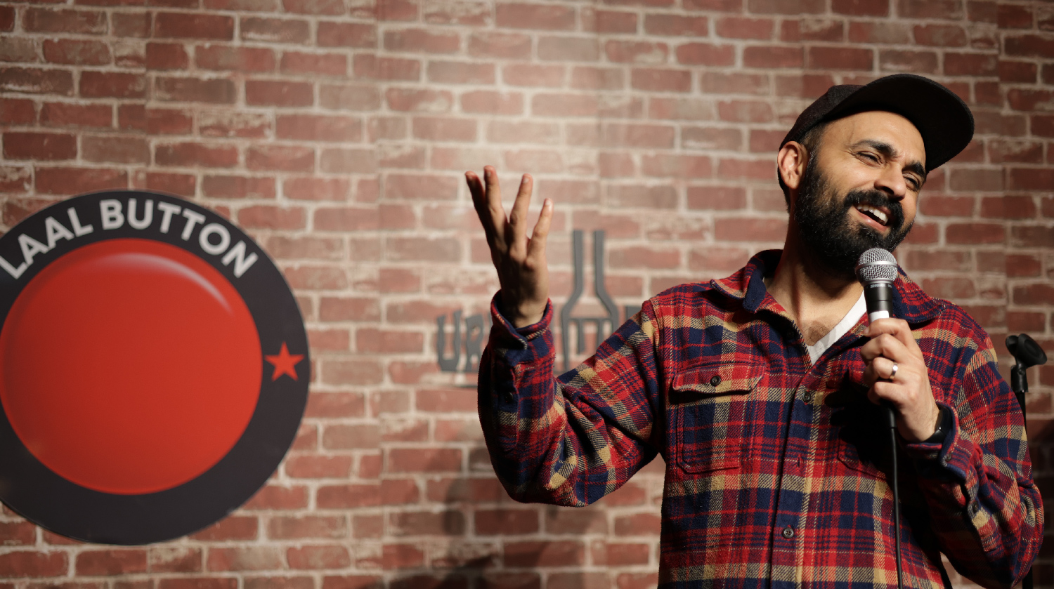 This image features a comedian or speaker performing on stage. The background displays a brick wall with a circular sign reading "Laal Button" and a microphone stand visible on the side. The performer is wearing a red-and-blue checkered flannel shirt and a cap, holding a microphone while gesturing expressively, suggesting they are mid-performance or delivering a humorous anecdote.