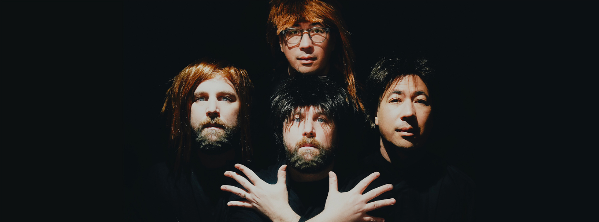 Four men posing dramatically in wigs with a dark background, parodying the iconic "Bohemian Rhapsody" Queen album cover.