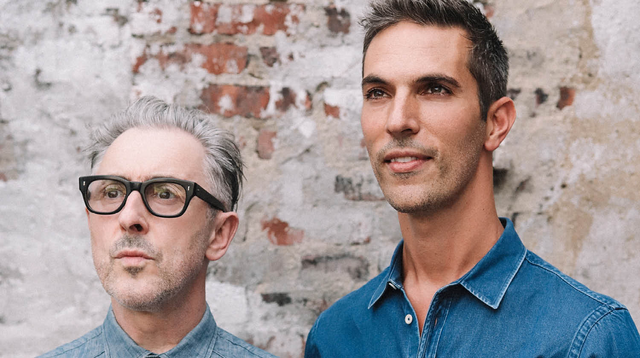 Alan Cumming and Ari Shapiro in denim, posed thoughtfully against a weathered brick wall. 