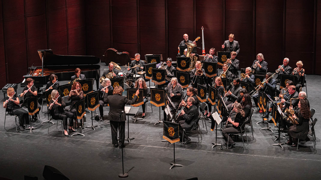 Full orchestra performing on stage with various brass, woodwind, and string instruments, and a grand piano.