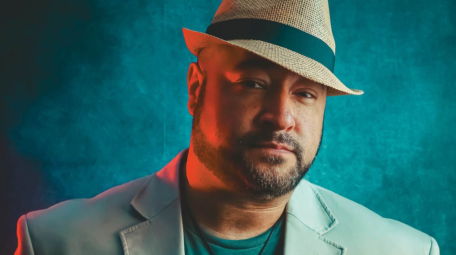 The image features a man wearing a light-colored jacket and a fedora hat with a dark band. His expression is serious and confident, with a subtle red light casting a warm hue on one side of his face, contrasting with the cool, blue-toned background.