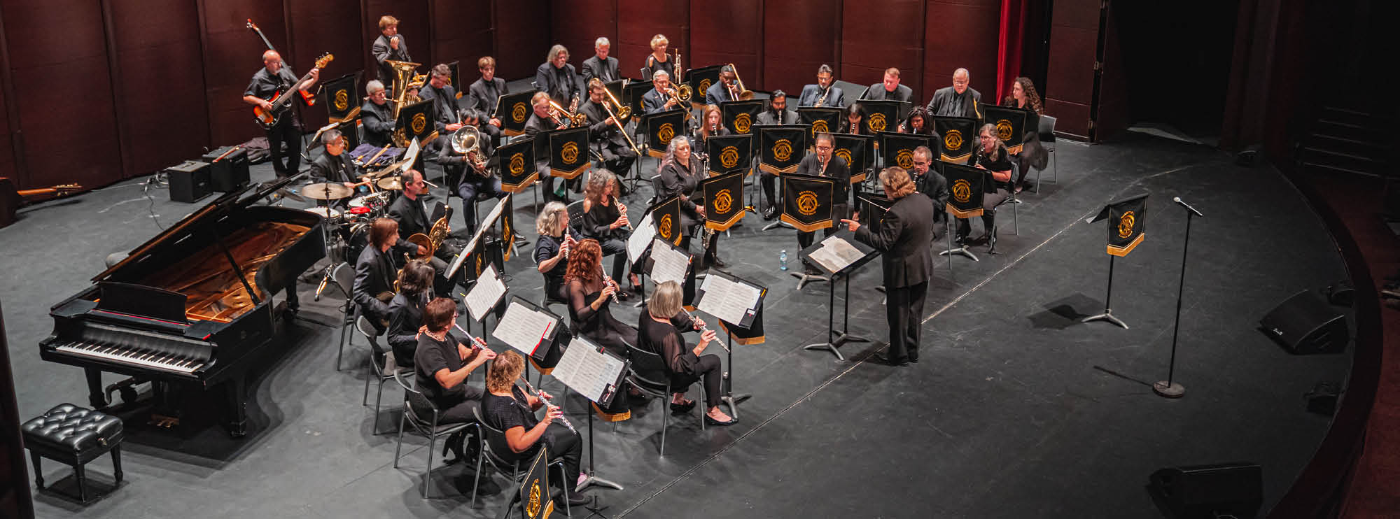 A full orchestra on stage with brass, strings, and a grand piano, music stands adorned with golden logos. 