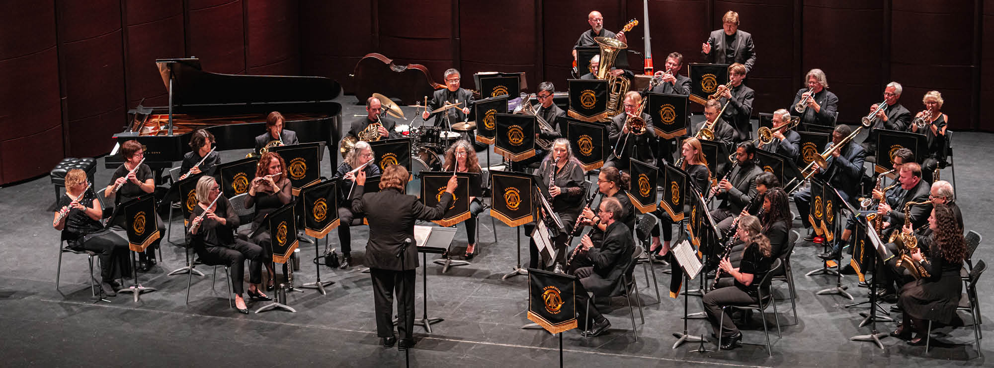 Full orchestra performing on stage with various brass, woodwind, and string instruments, and a grand piano.