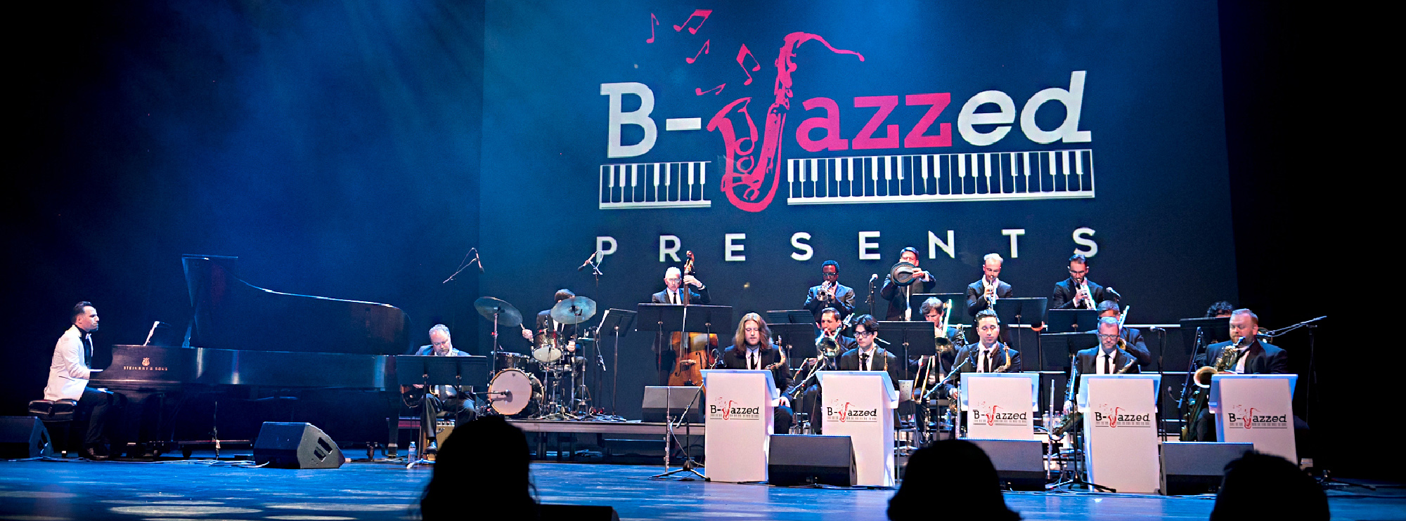 A jazz band performs on stage at the "B-Jazzed Presents" event. The large ensemble includes musicians playing various instruments such as the piano, drums, trumpets, trombones, and saxophones. The stage is bathed in blue lighting, and the backdrop features a large projection of the "B-Jazzed" logo, which includes a stylized saxophone and piano keys.