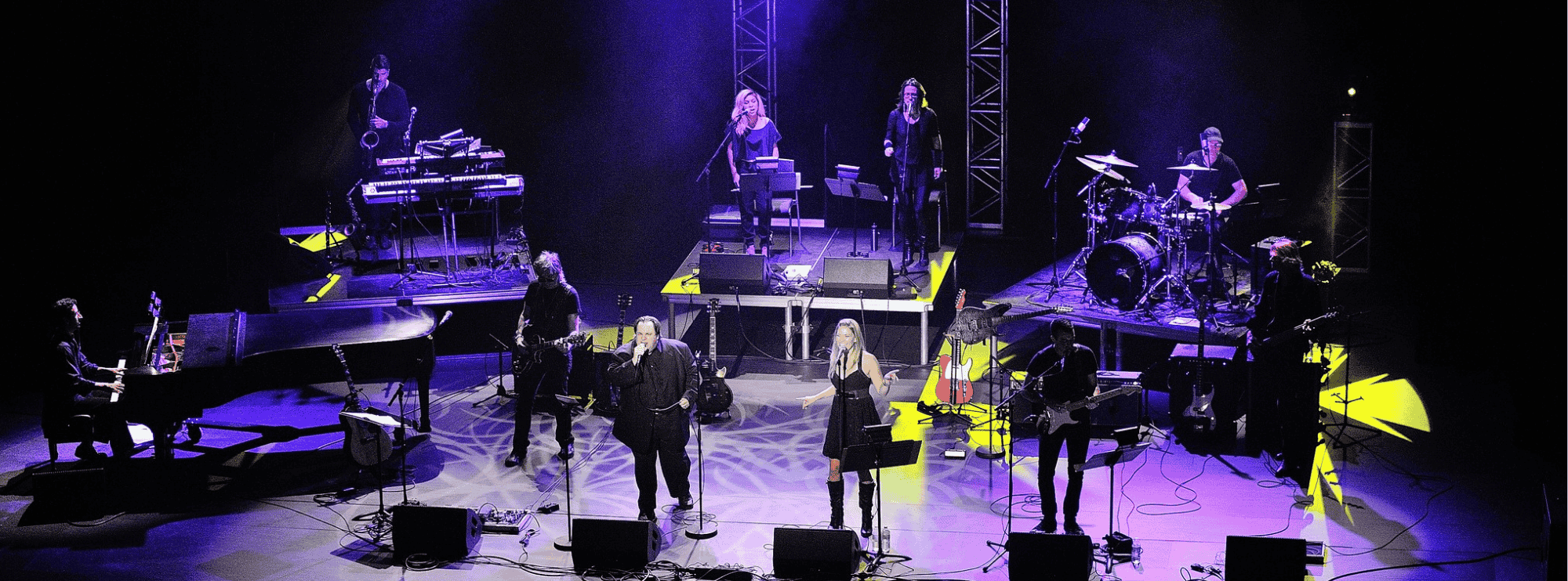 The image captures a band performing on stage, surrounded by speakers, microphones, and wires. Singers take center stage, while musicians play drums, guitar, and keyboard. Colorful lighting illuminates the stage floor.