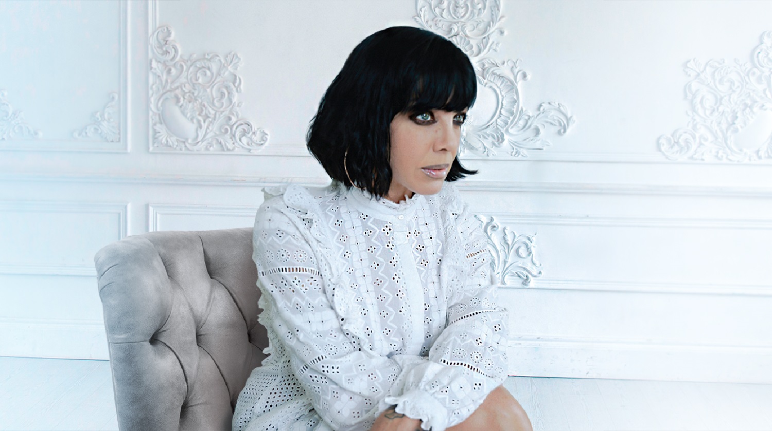 A stylish woman sits on a plush, light-colored chair in an elegantly decorated room. She has a chic bob haircut and wears a white, intricately patterned lace dress. The background features detailed, ornate wall panels.