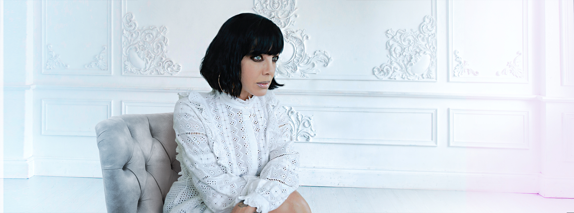 A stylish woman sits on a plush, light-colored chair in an elegantly decorated room. She has a chic bob haircut and wears a white, intricately patterned lace dress. The background features detailed, ornate wall panels.