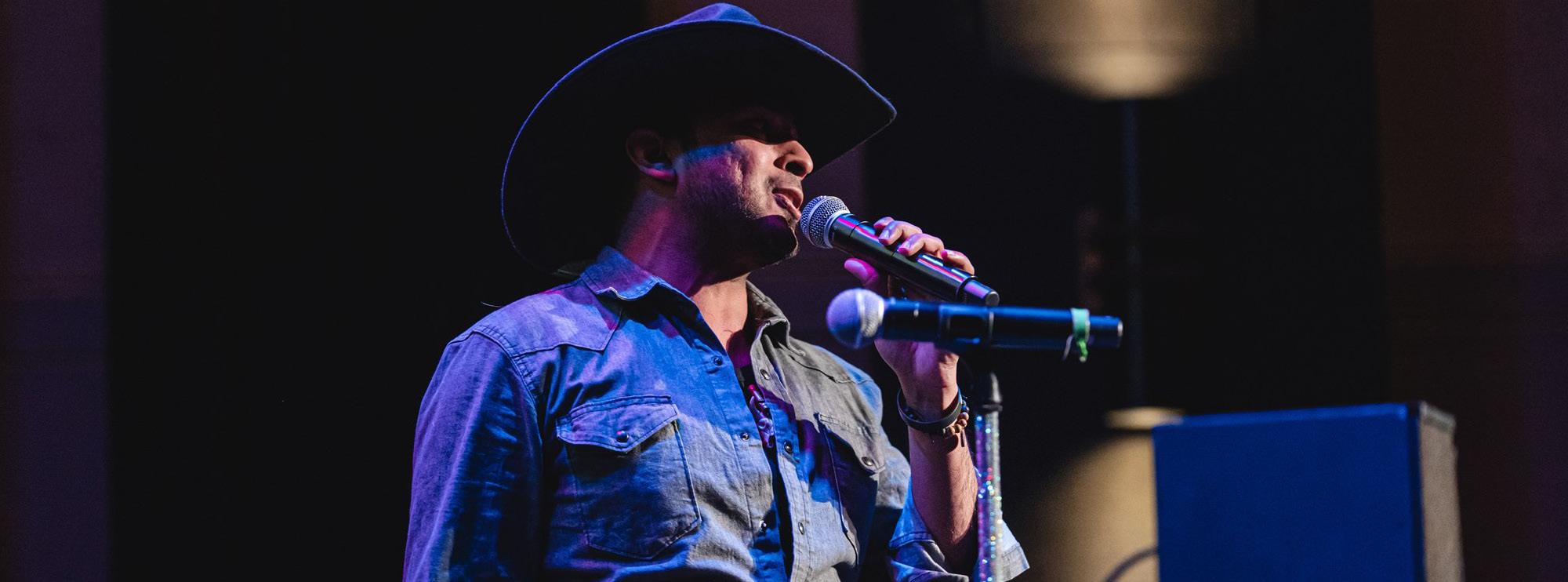 The image captures a male performer wearing a black cowboy hat and a denim shirt, singing into a microphone on stage. The stage is dimly lit with a deep blue and purple ambiance, casting shadows on the background. The performer holds the microphone stand with one hand, and a large speaker and a faint stage light are visible in the background.