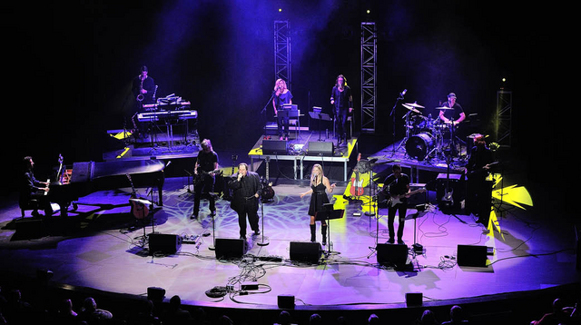 The image captures a band performing on stage, surrounded by speakers, microphones, and wires. Singers take center stage, while musicians play drums, guitar, and keyboard. Colorful lighting illuminates the stage floor.