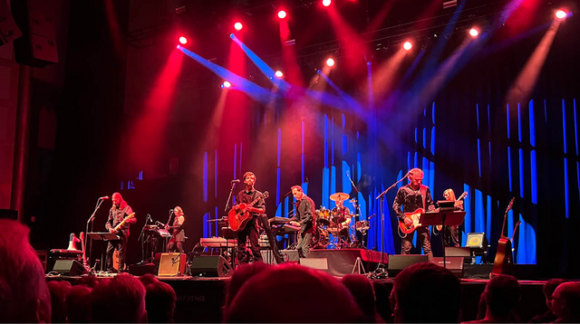 Image of several musicians performing on stage, playing an array of instruments including the guitar, drums, and piano. Speakers, music stands, and wires are scattered across the stage, while blue and red lights illuminate the space. The tops of heads can be seen as the audience observes from below.